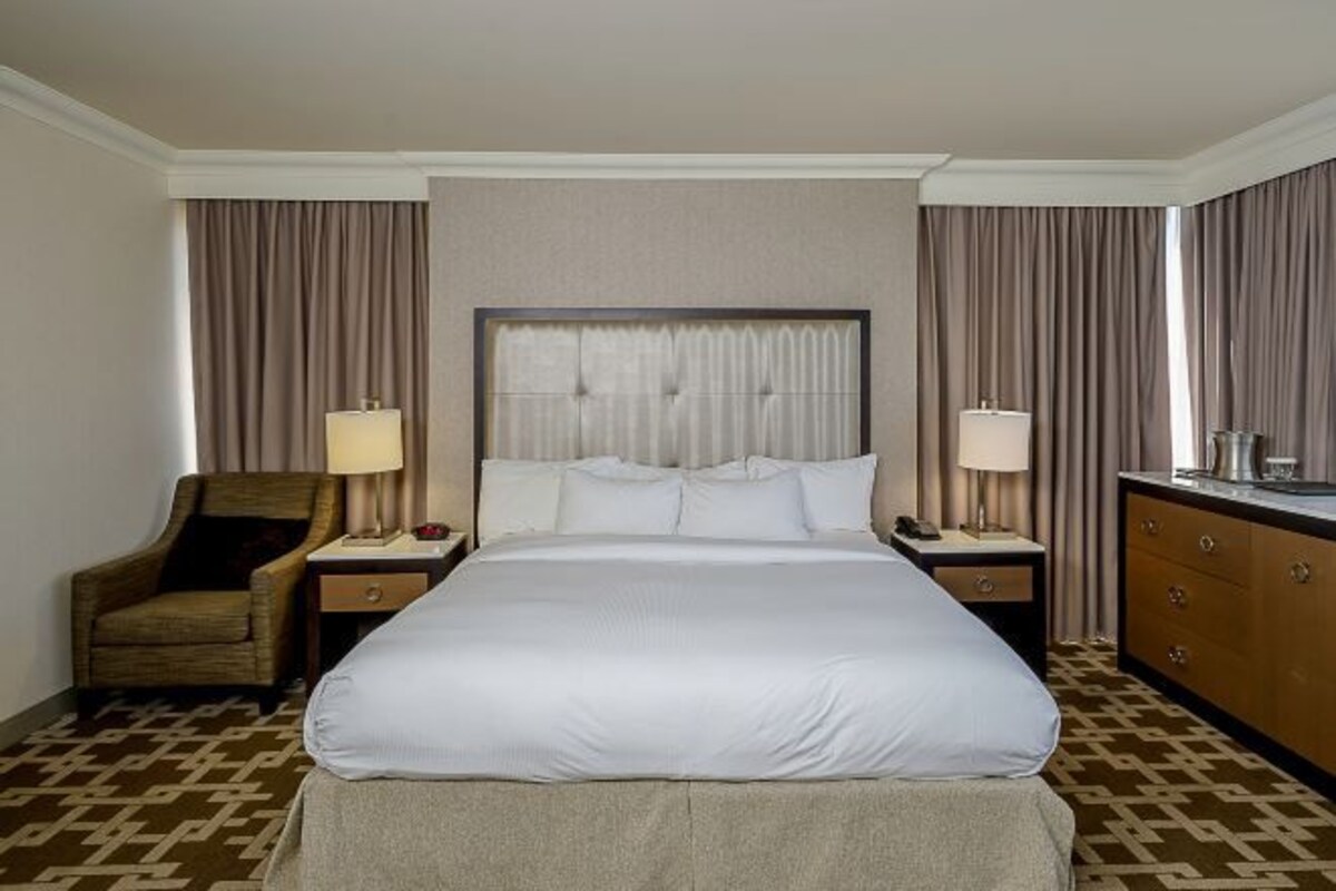 Une chambre d’hôtel avec un grand lit, deux tables de chevet avec des lampes, une commode en bois et une moquette à motifs.