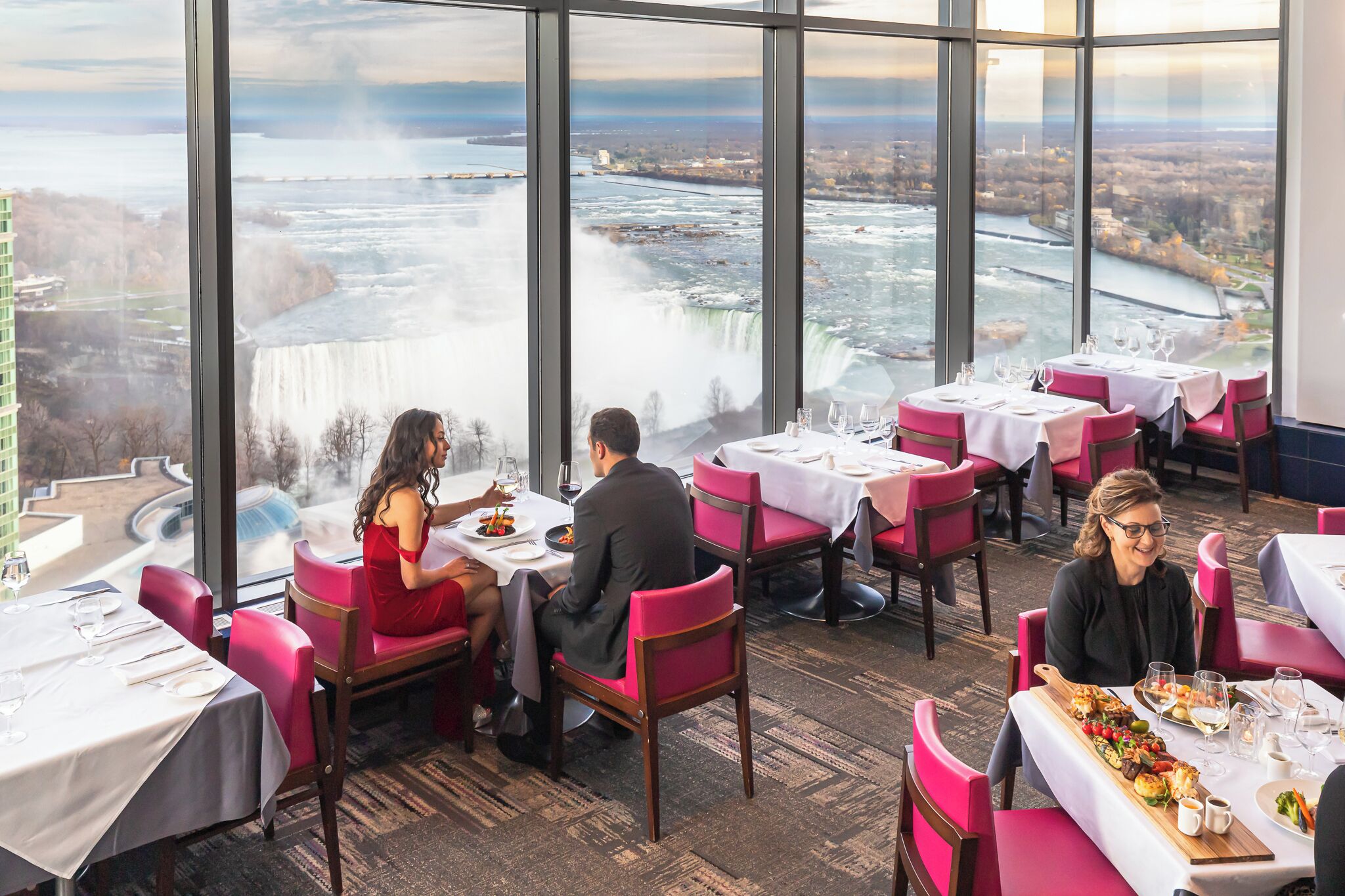 Un restaurante con mesas dispuestas para comer, amplias ventanas que ofrecen vistas a una cascada y un río, y un paisaje urbano al fondo.