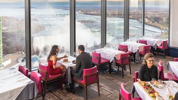Un restaurante con mesas dispuestas para comer, amplias ventanas que ofrecen vistas a una cascada y un río, y un paisaje urbano al fondo.