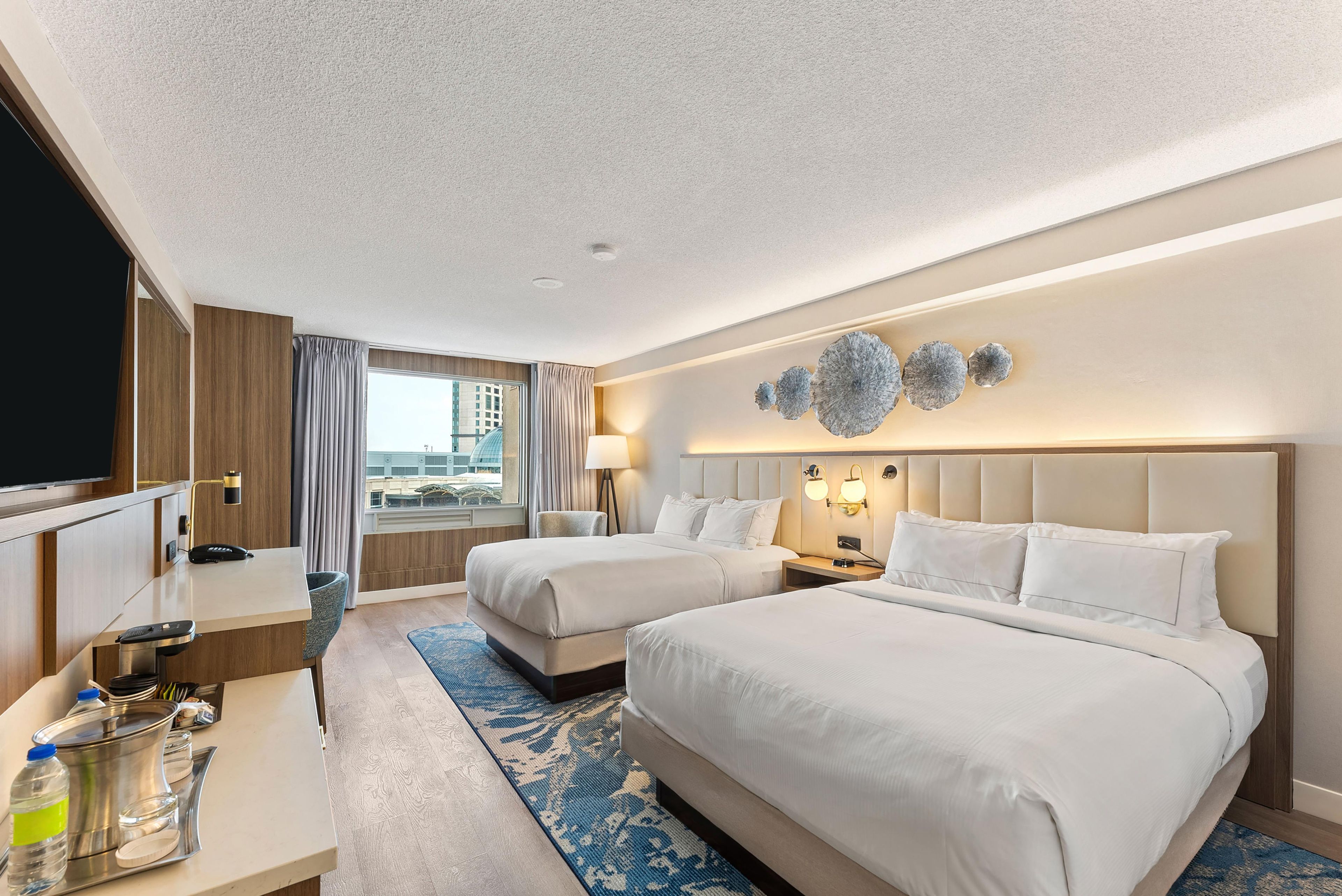 Quarto de hotel com duas camas, uma escrivaninha, televisão e janela com cortinas.