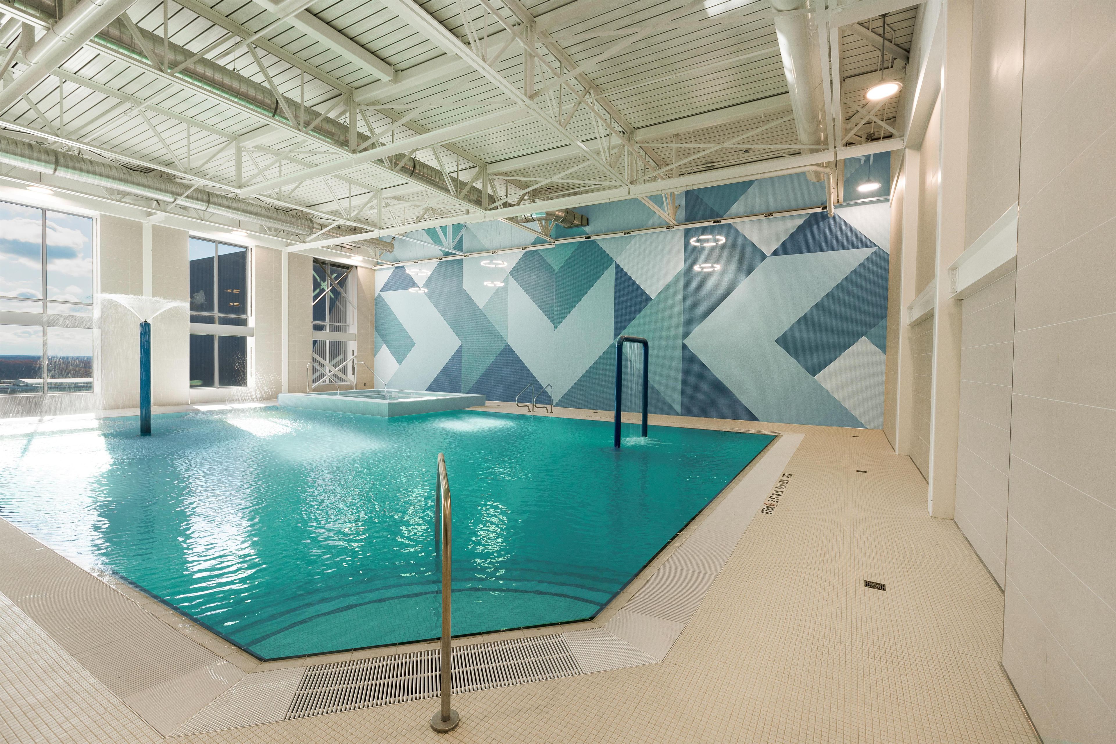 An indoor swimming pool with a geometric wall design, a high ceiling with visible beams, and a clear view of the sky through large windows.