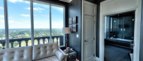 A modern living room with a large window, a sofa, a small table with a lamp, and a door leading to a balcony with a city view.