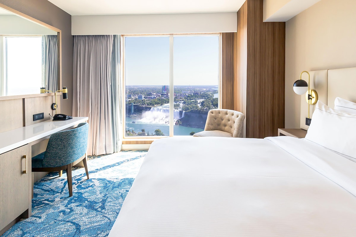 A hotel room with a large bed, a desk, a chair, a nightstand, and a view of a waterfall.