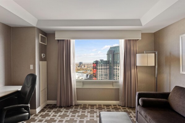 A hotel room with a large window, a desk, a chair, a sofa, and a lamp.