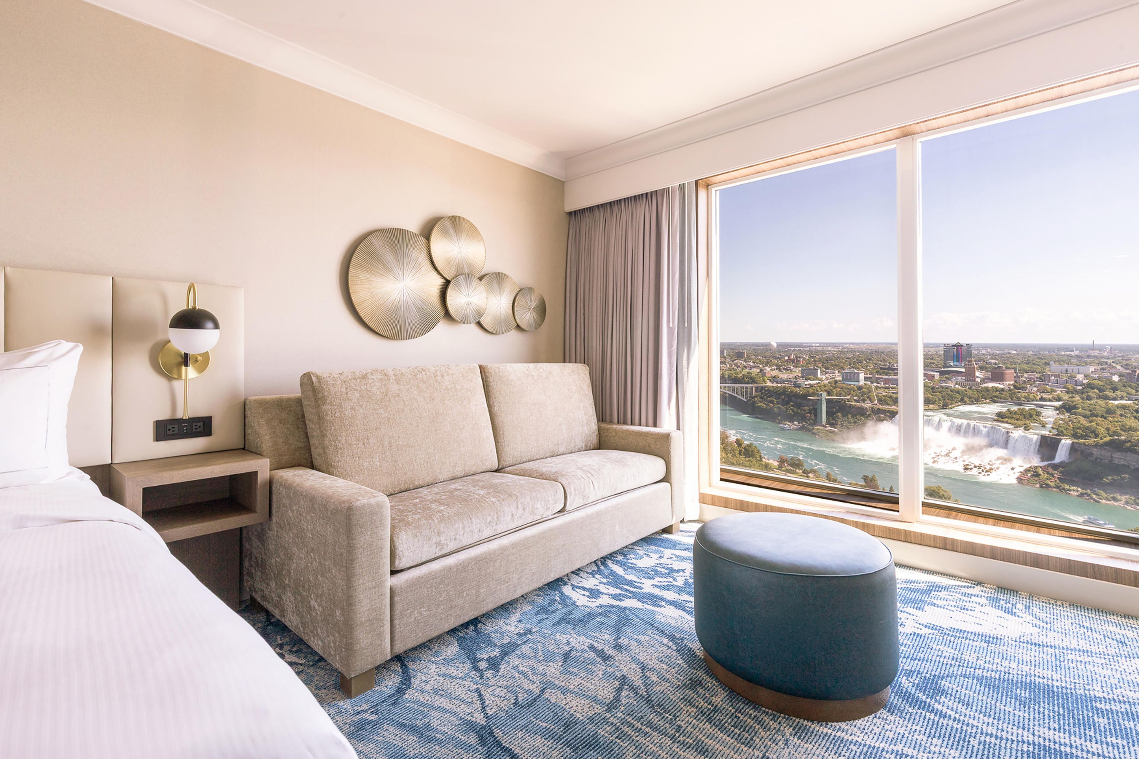 Una camera d'albergo con un letto, una poltrona, un poggiapiedi blu, una lampada a parete e un'ampia finestra con vista sulla città e sul fiume.