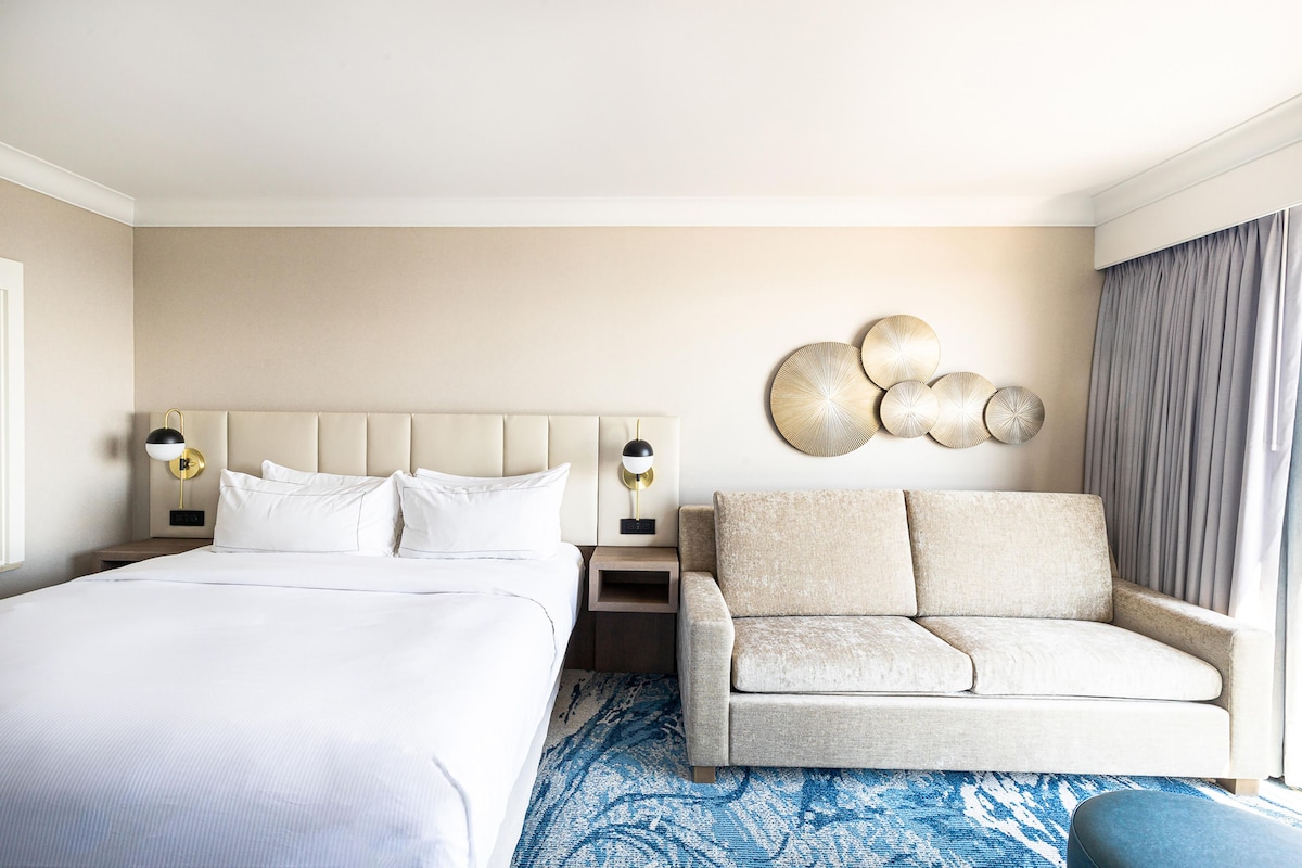 A hotel room with a large bed, a sofa, a wall-mounted decorative piece, and a window with curtains.