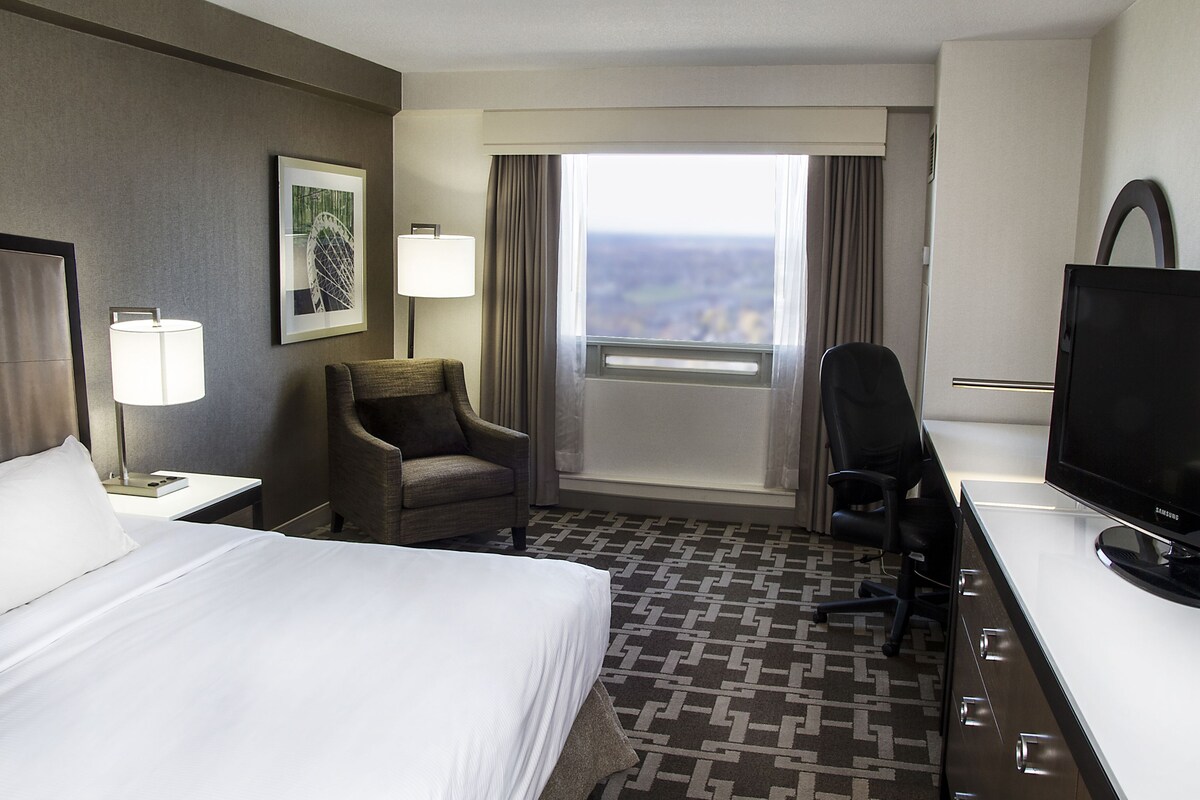 A hotel room with a bed, a desk, a chair, a TV, a window with curtains, and a lamp.
