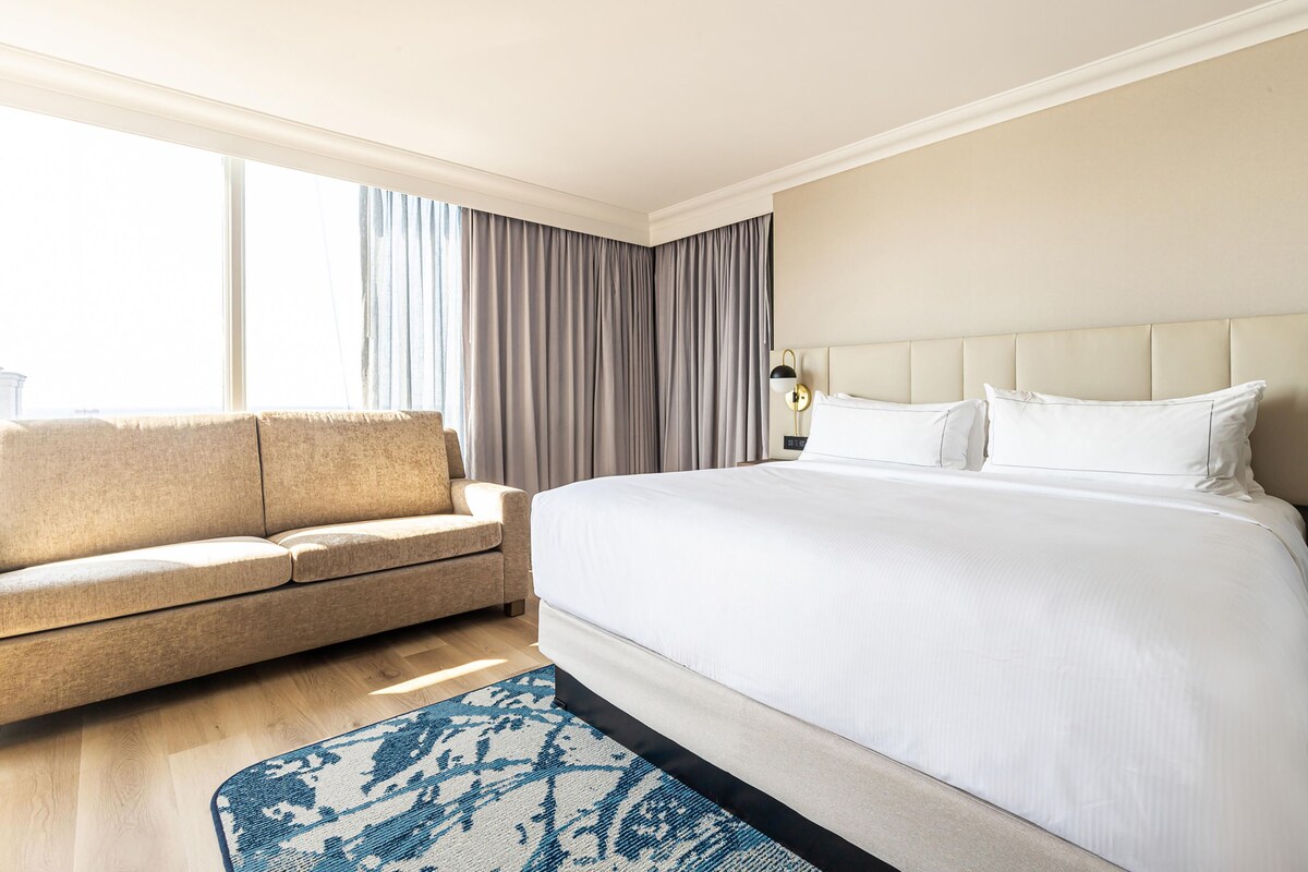 A hotel room with a large bed, a sofa, a nightstand, and a window with curtains.