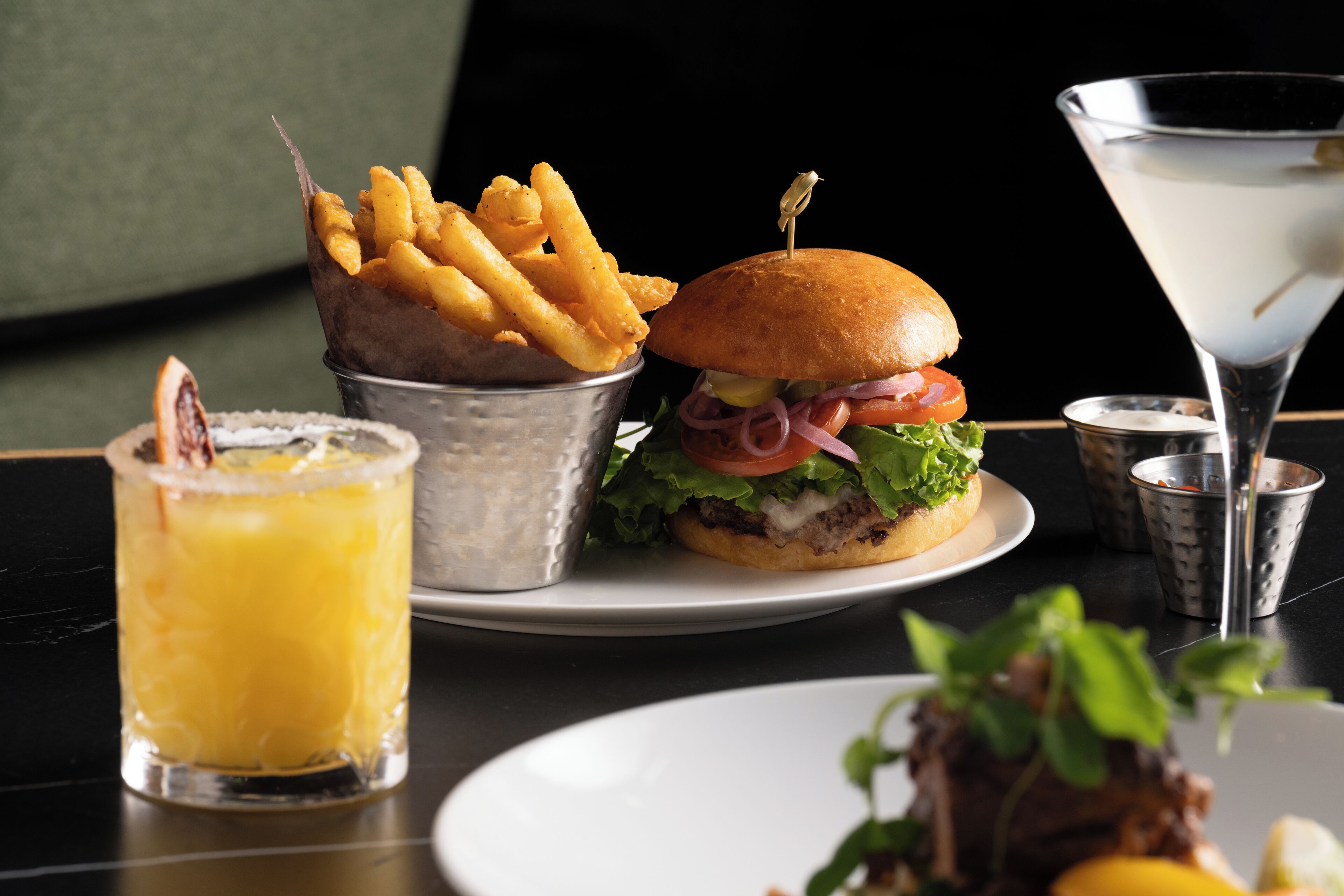 A meal consisting of a burger, fries, a martini, and a drink on a table.