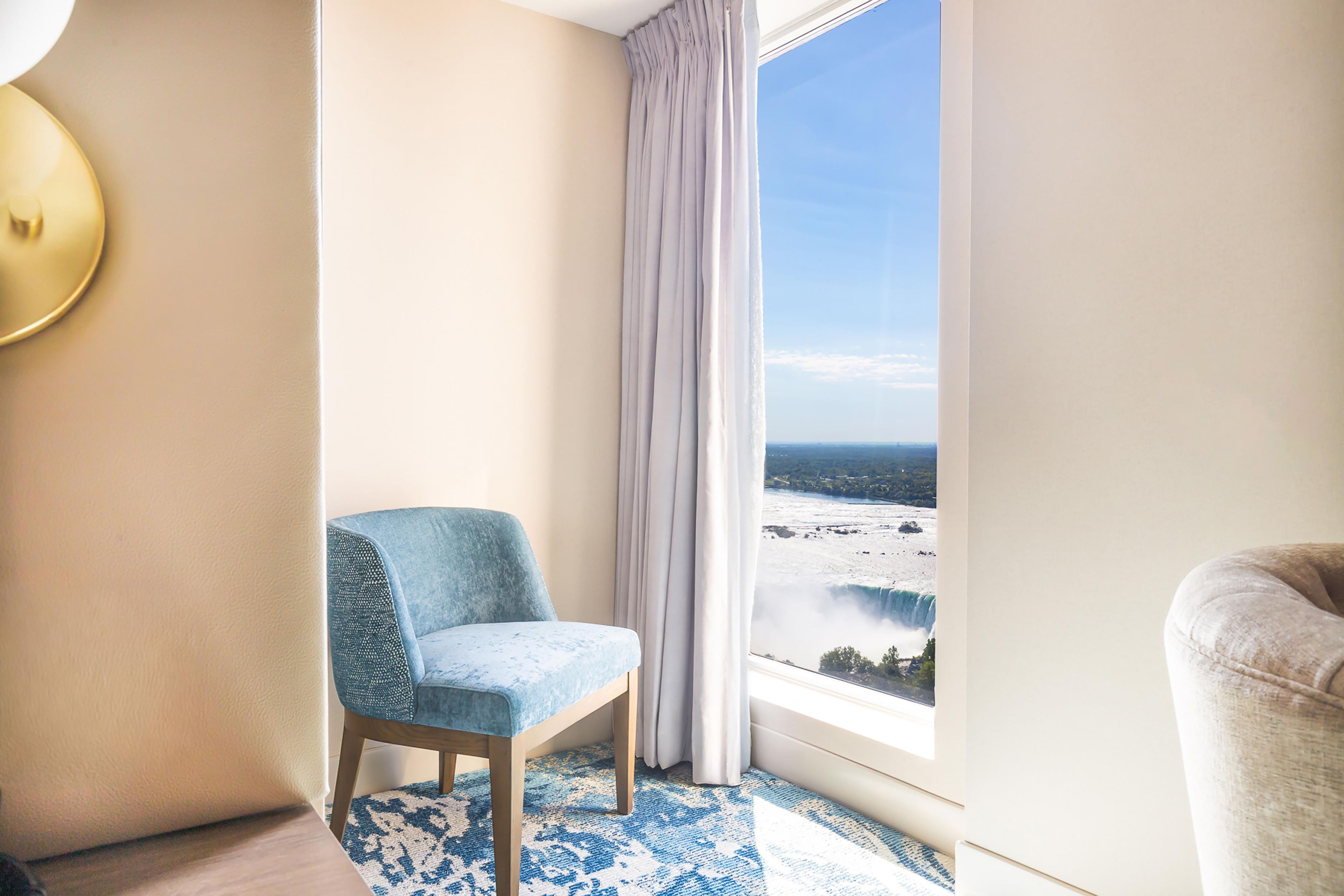 Habitación de hotel con vista a una cascada, un sillón azul, un tapete estampado y un amplio ventanal con cortinas.
