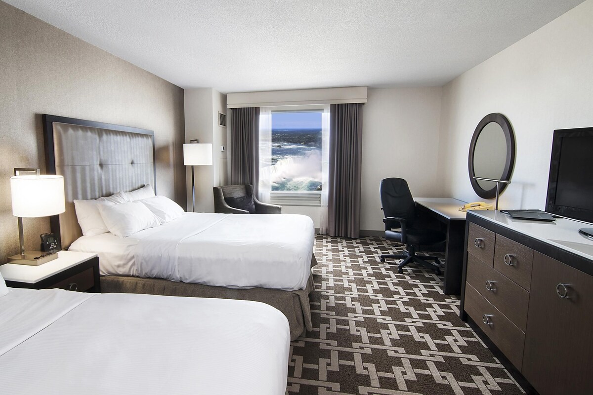 A hotel room with two beds, a desk with a computer, a chair, a TV, and a view of the ocean.