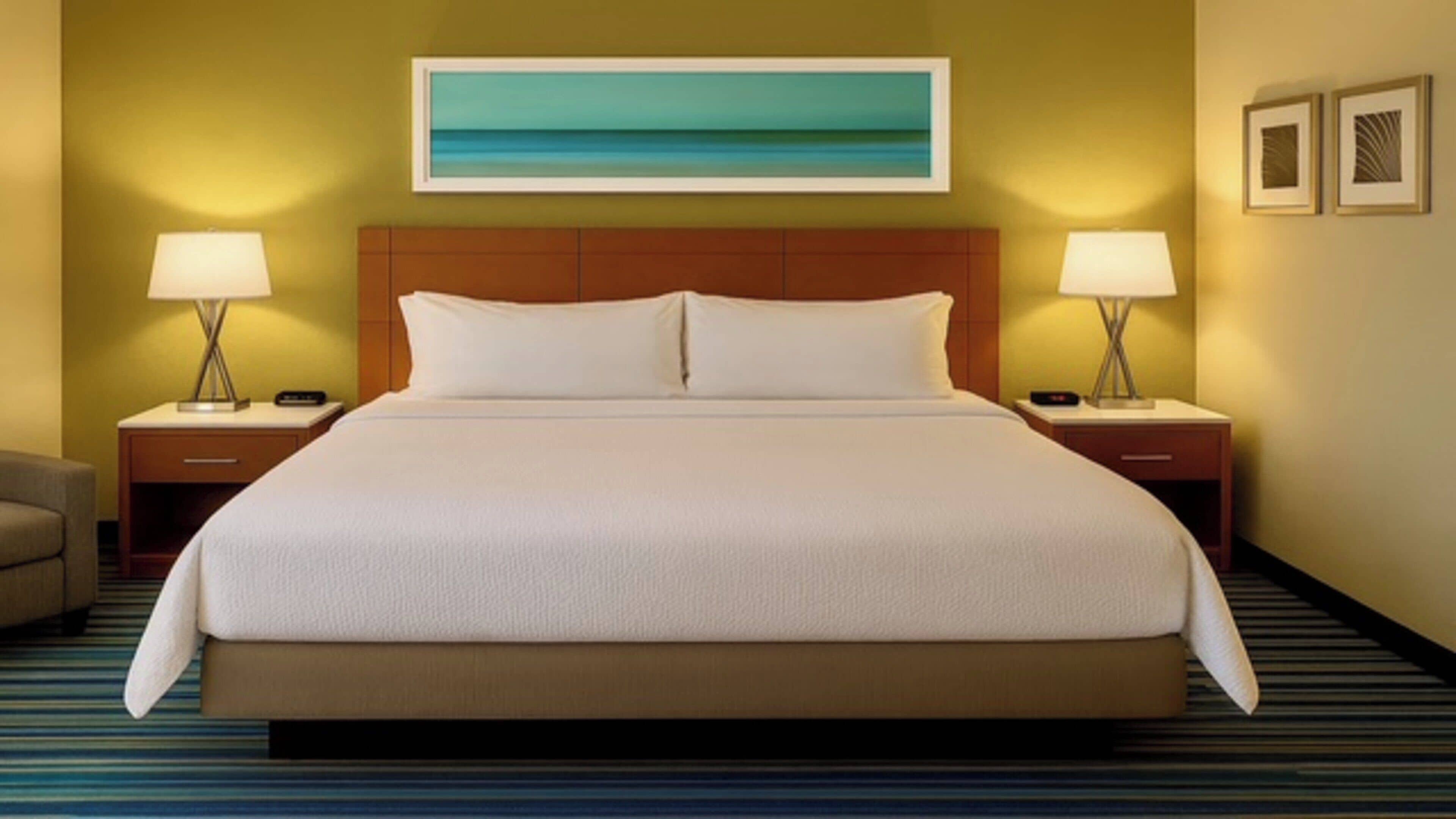 A hotel room with a large bed, two bedside tables with lamps, a headboard, a painting on the wall, and a striped carpet.