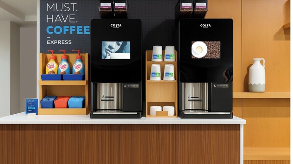 Eine Kaffeestation mit drei Kaffeeautomaten, beschriftet als 'Costa Coffee Express', und einer Auswahl an Kaffeezutaten, darunter 'Coffee Mate'-Flaschen und Einwegbechern.