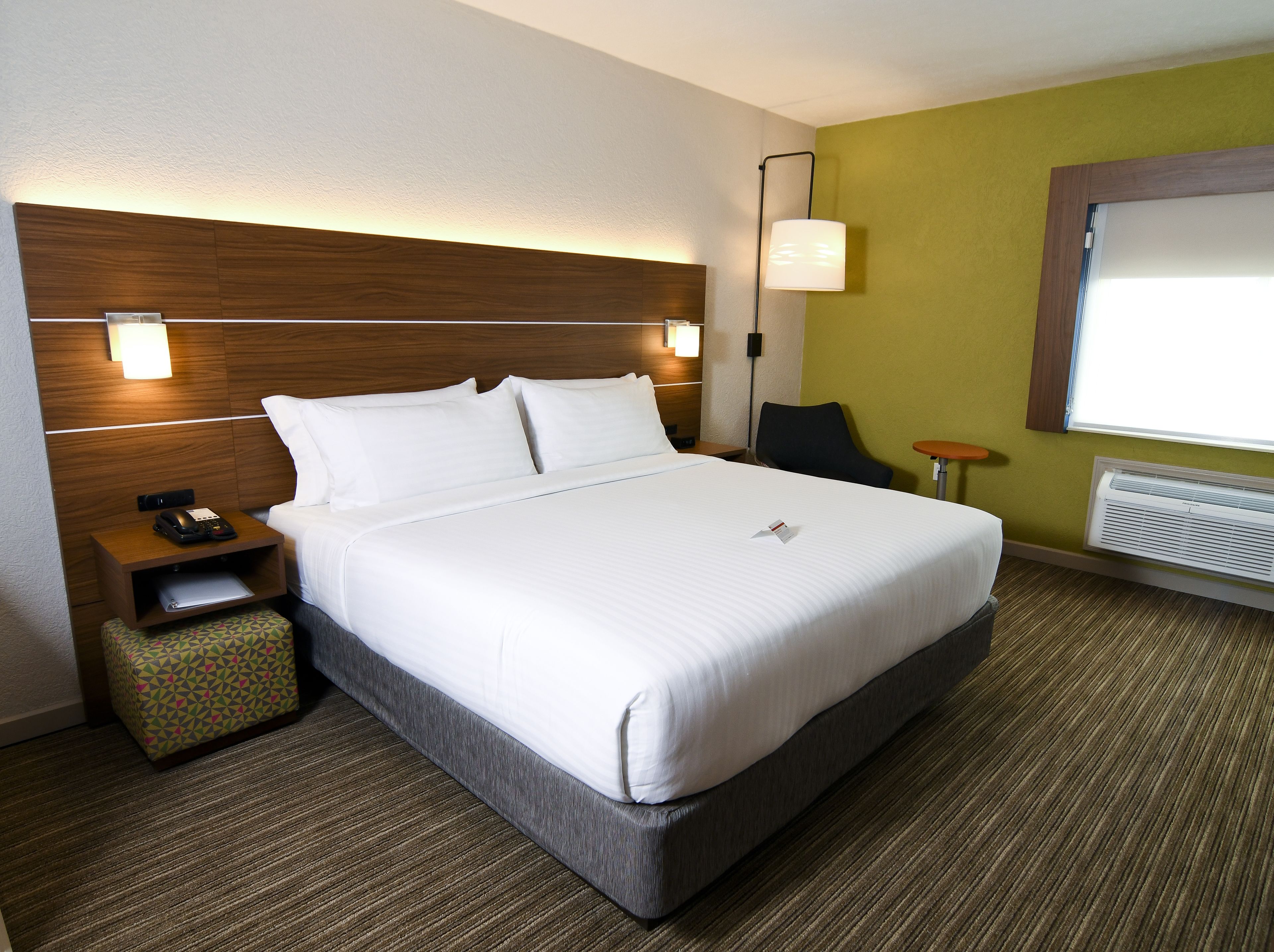 Habitación de hotel con una cama grande, una mesita de noche con teléfono, un taburete pequeño, una silla, una lámpara de pared y una ventana con unidad de aire acondicionado.