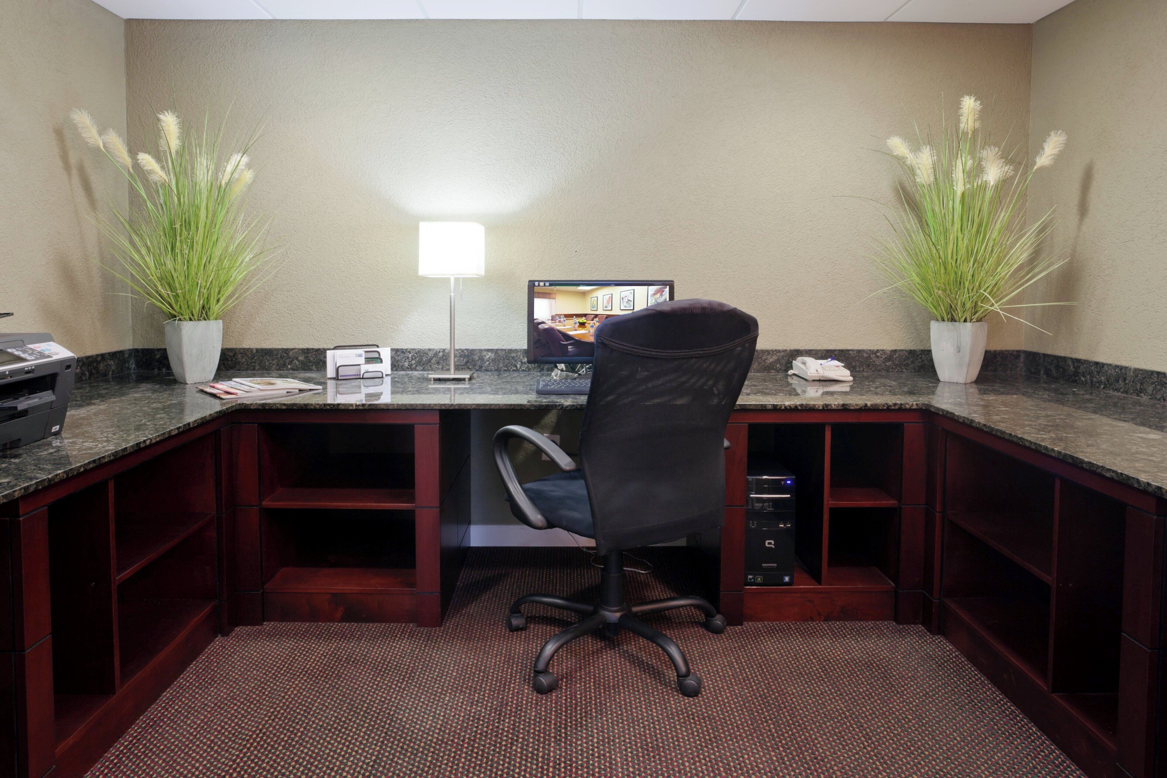 A hotel office with a desk, chair, two potted plants, a lamp, and a computer monitor.