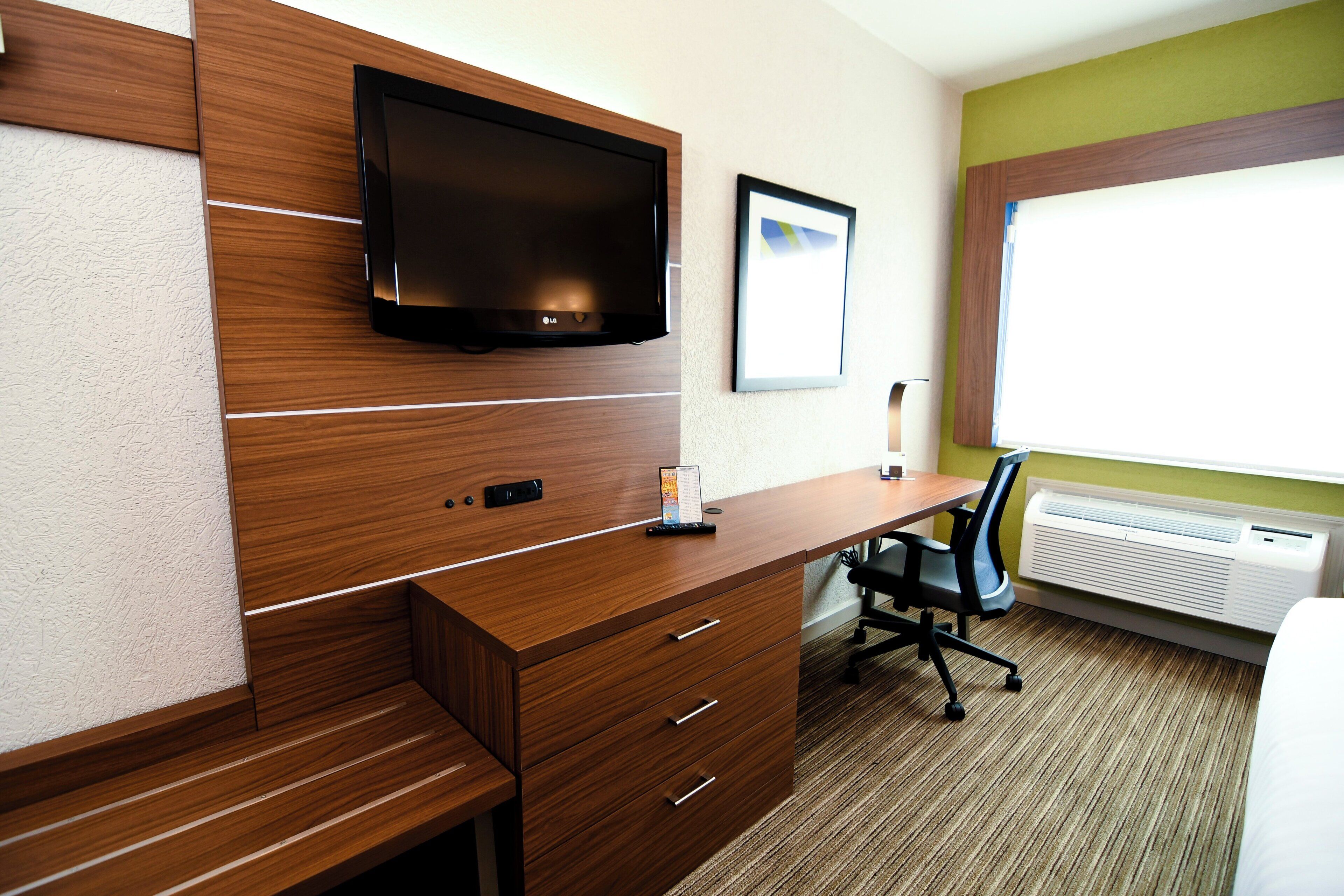 A hotel room with a wooden desk, a flat-screen TV mounted on the wall, a framed picture, and an air conditioning unit.