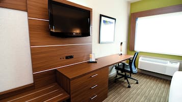 A hotel room with a wooden desk, a flat-screen TV mounted on the wall, a framed picture, and an air conditioning unit.