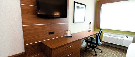 A hotel room with a wooden desk, a flat-screen TV mounted on the wall, a framed picture, and an air conditioning unit.