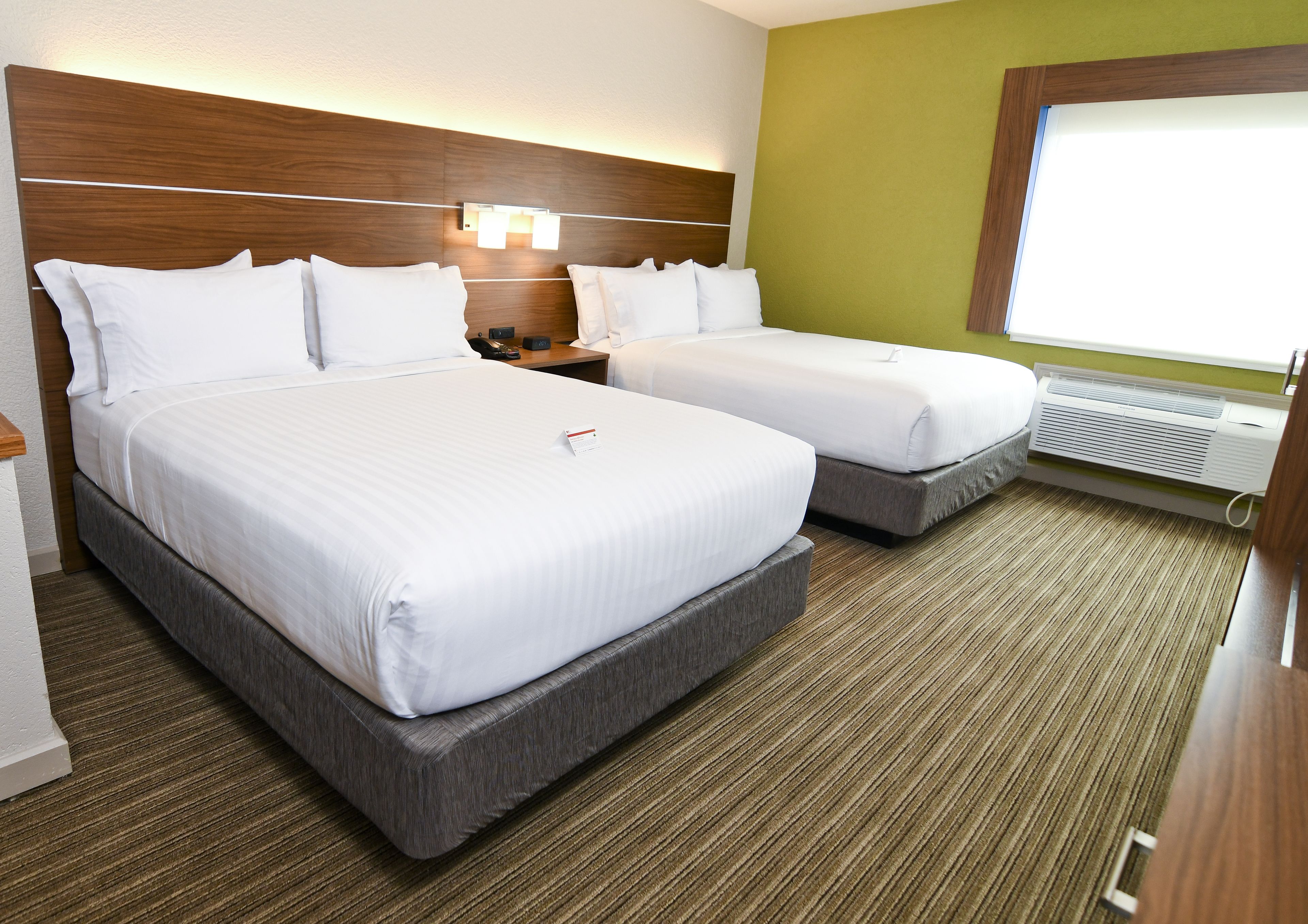 A hotel room with two beds, a wooden headboard, a nightstand, a bedside table, a window with blinds, and a wall-mounted air conditioning unit.