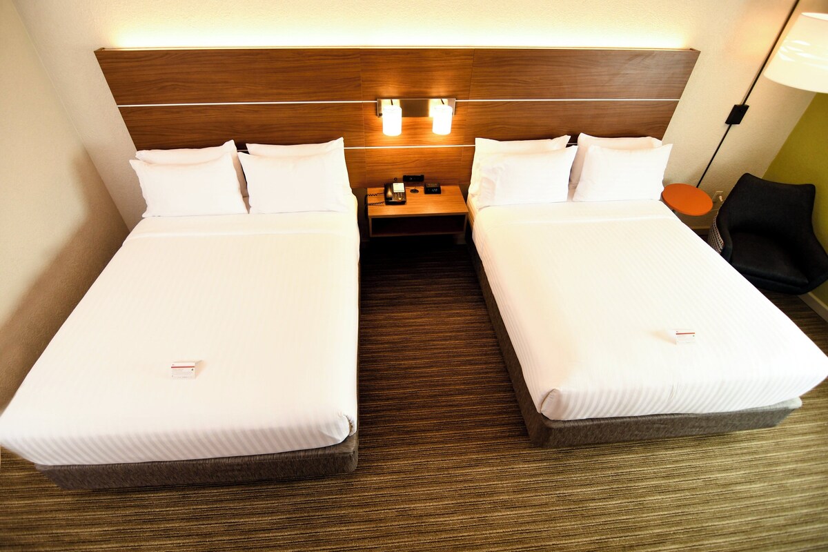 Habitación de hotel con dos camas, cabecero de madera, lámparas de noche, mesitas de noche y una silla.