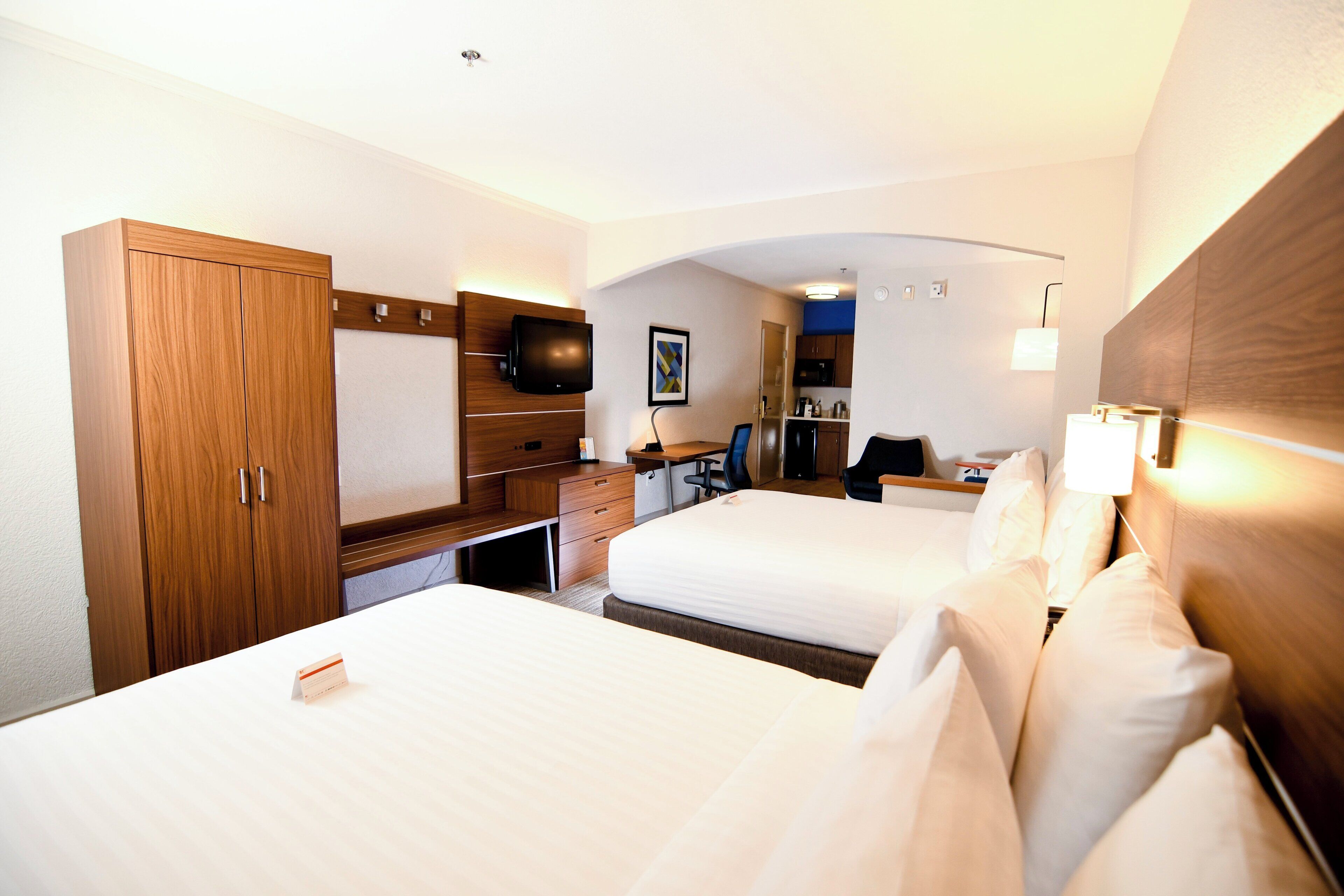 Quarto de hotel com duas camas, uma mesa, uma cadeira, televisão e uma pequena cozinha.