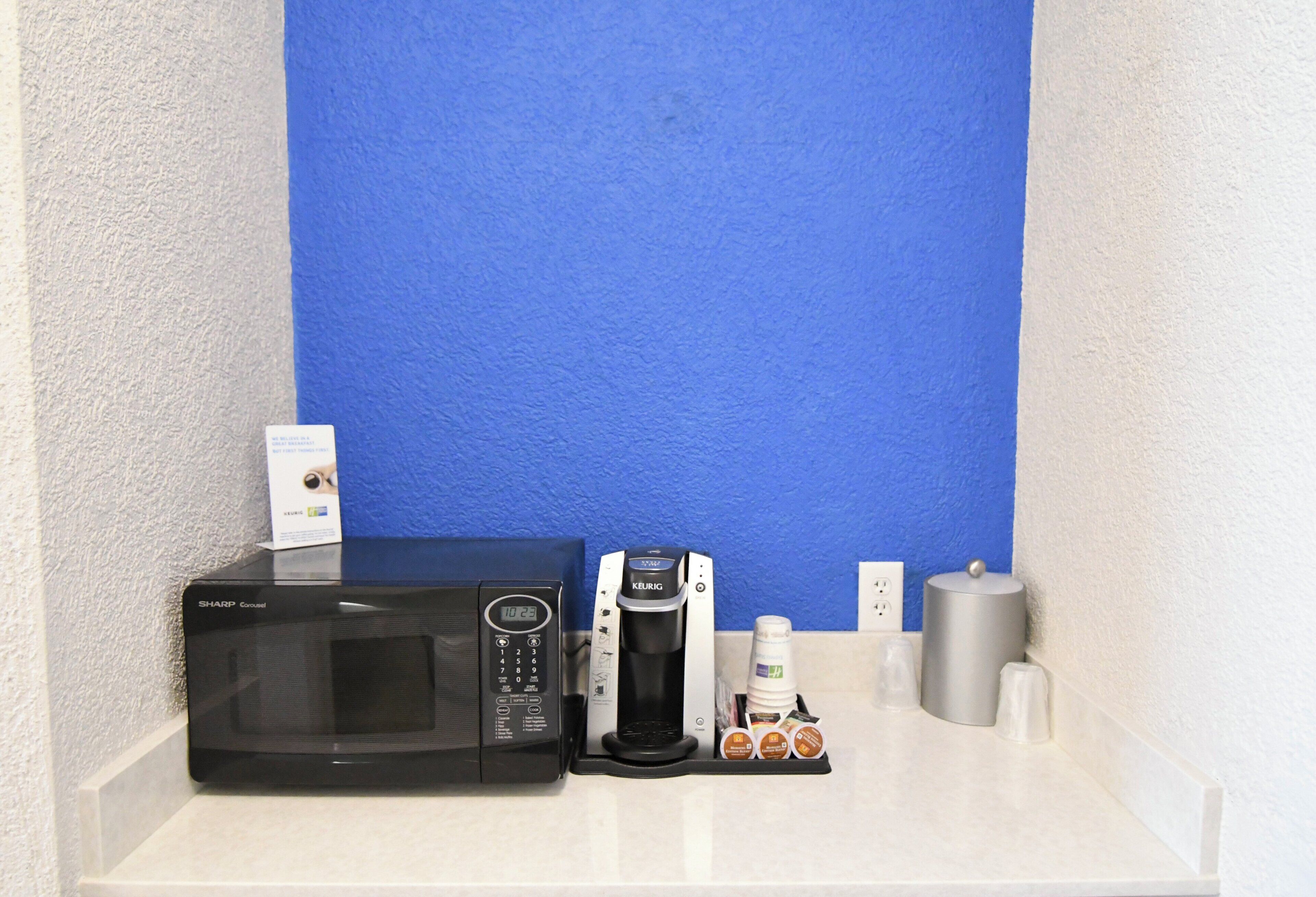 Una kitchenette con microondas, cafetera y cápsulas de café sobre una encimera blanca.