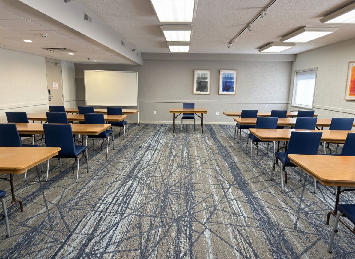 Un salón de conferencias con piso alfombrado, mesas, sillas, una pantalla de proyector y cuadros en la pared.