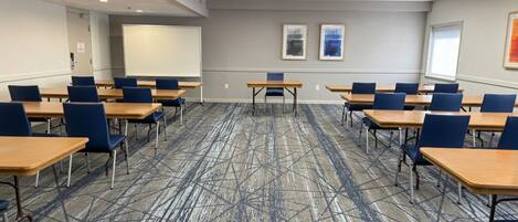 Un salón de conferencias con piso alfombrado, mesas, sillas, una pantalla de proyector y cuadros en la pared.