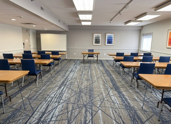 Un salón de conferencias con piso alfombrado, mesas, sillas, una pantalla de proyector y cuadros en la pared.