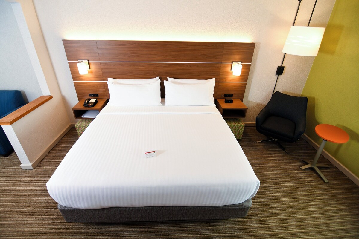 Una habitación de hotel con una cama grande, mesitas de noche, una silla, una lámpara y una pequeña mesa.