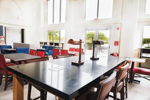 Uma área de refeições moderna com pé direito alto, grandes janelas e diferentes opções de assentos, incluindo mesas e cadeiras.