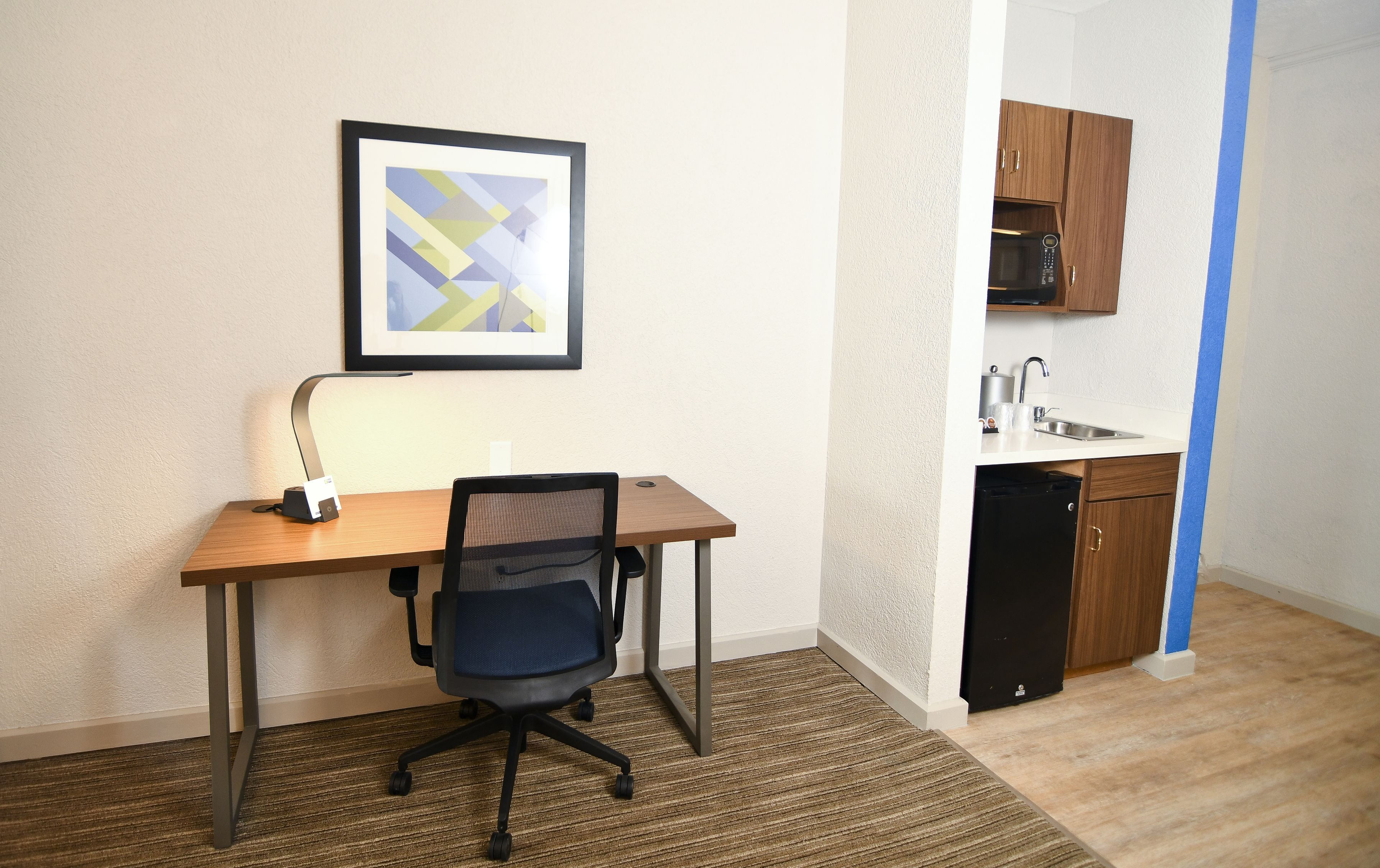 A small room with a wooden desk, a black office chair, a framed abstract painting on the wall, and a kitchenette with wooden cabinets and a microwave.