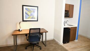A small room with a wooden desk, a black office chair, a framed abstract painting on the wall, and a kitchenette with wooden cabinets and a microwave.