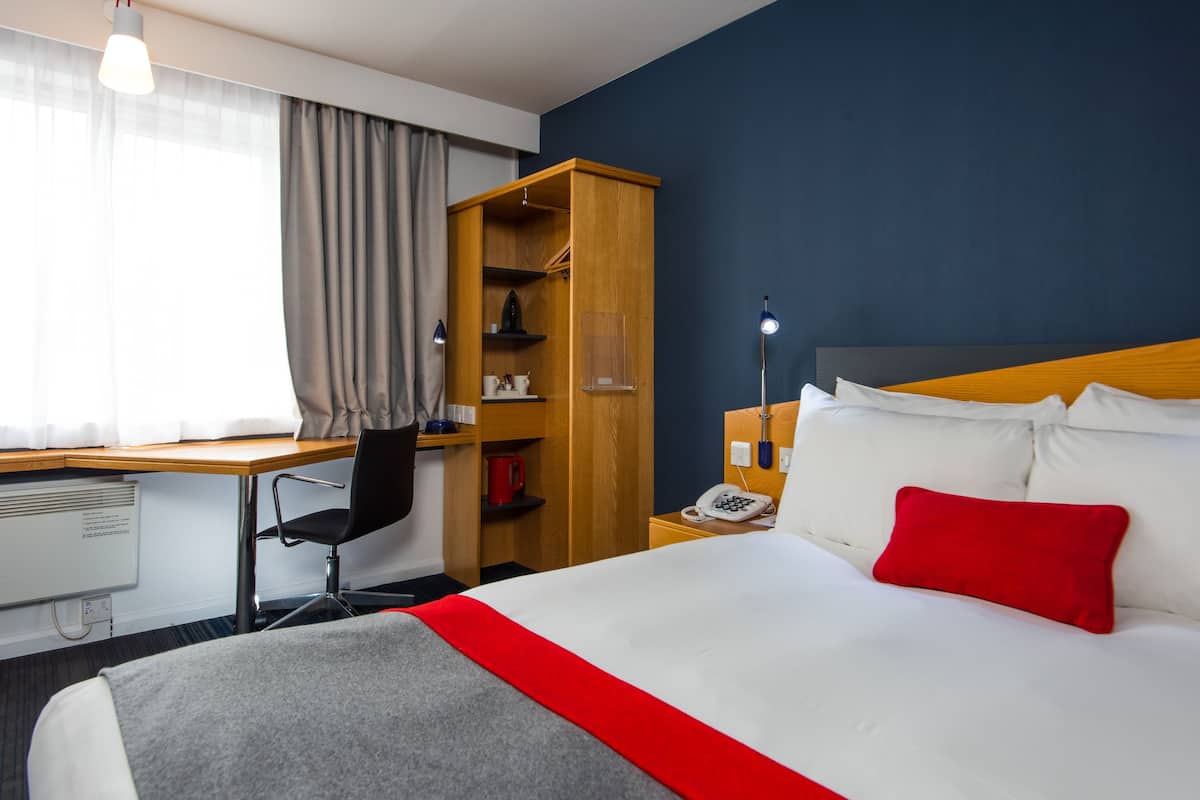 Habitación de hotel con cama, escritorio, silla, armario y mesita de noche con lámpara.