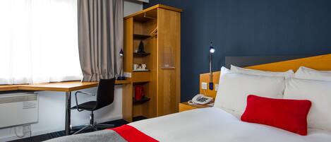 Ein Hotelzimmer mit einem Bett, einem Schreibtisch, einem Stuhl, einem Kleiderschrank und einem Nachttisch mit Lampe.