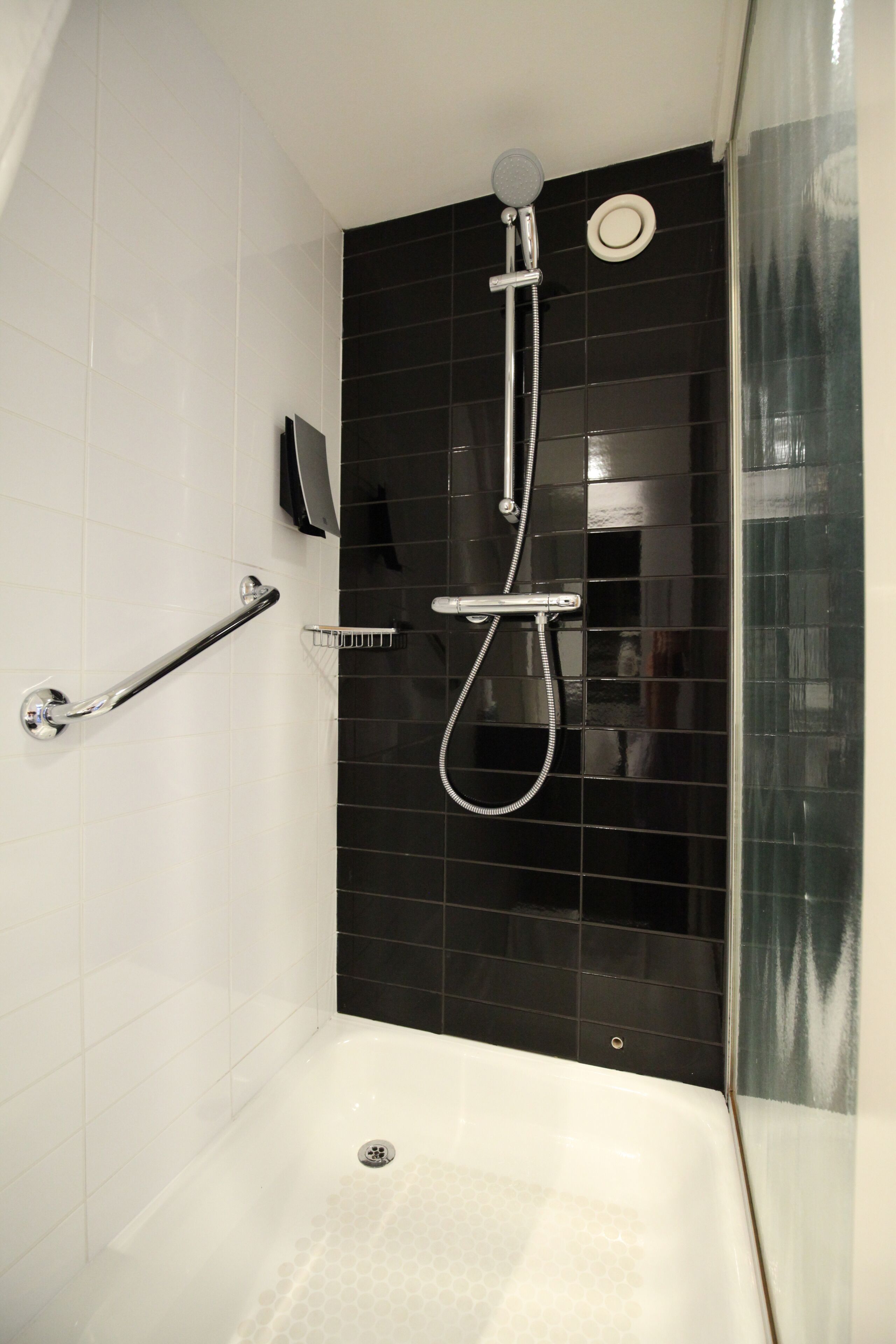 A modern bathroom with a black tiled shower wall, a white bathtub, and a handrail.