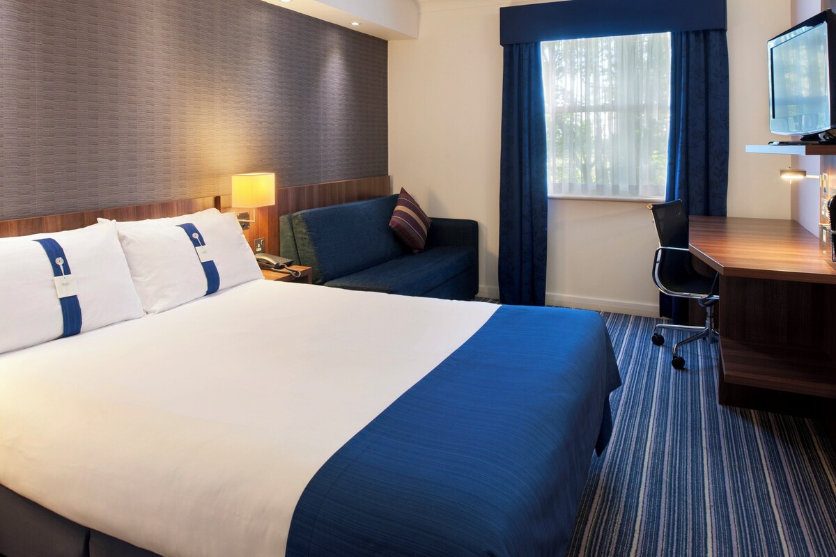 A hotel room with a large bed, a desk with a chair, a television, and a window with curtains.