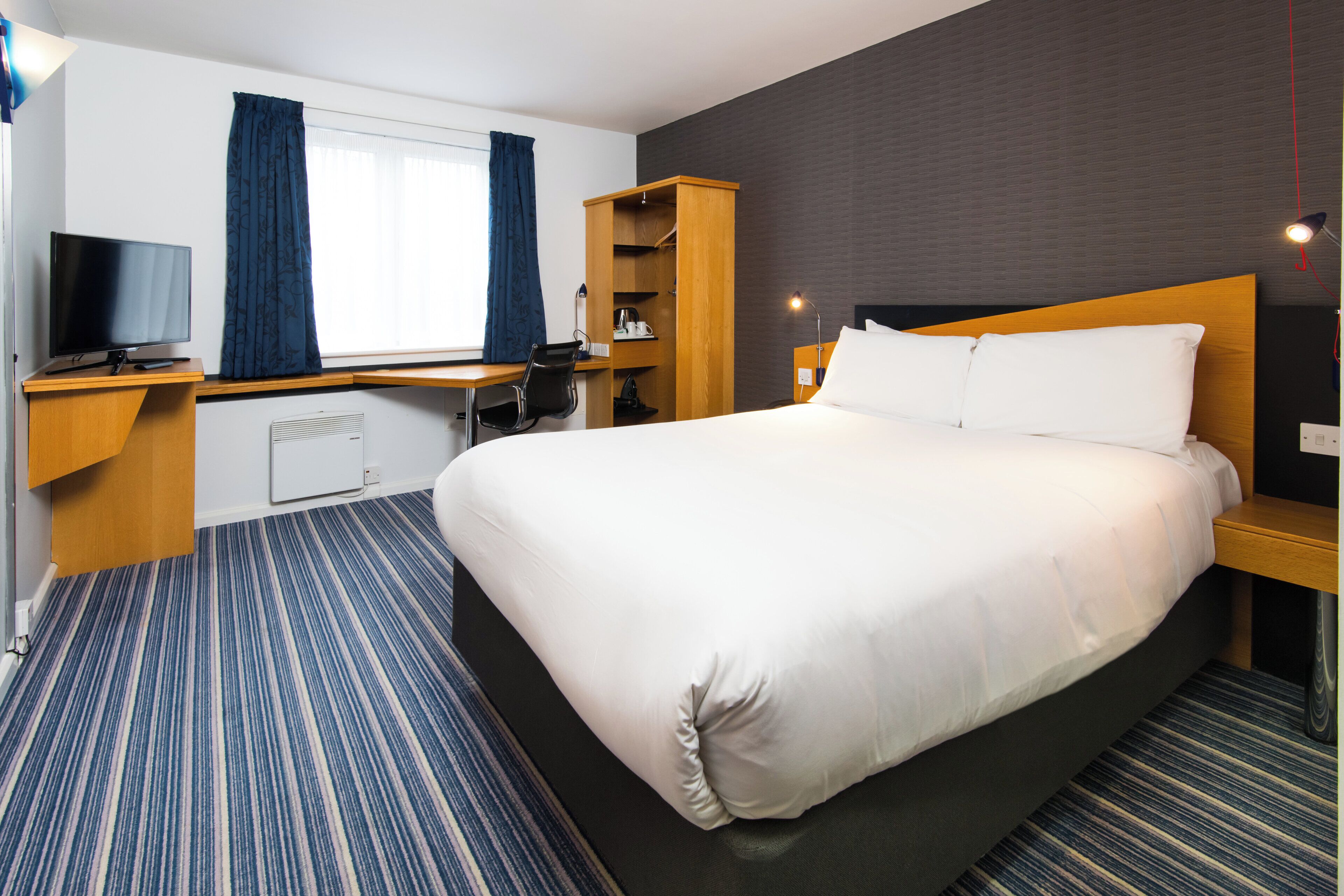 Ett hotellrum med en stor säng, ett skrivbord med en tv, en stol, en garderob och ett fönster med gardiner.