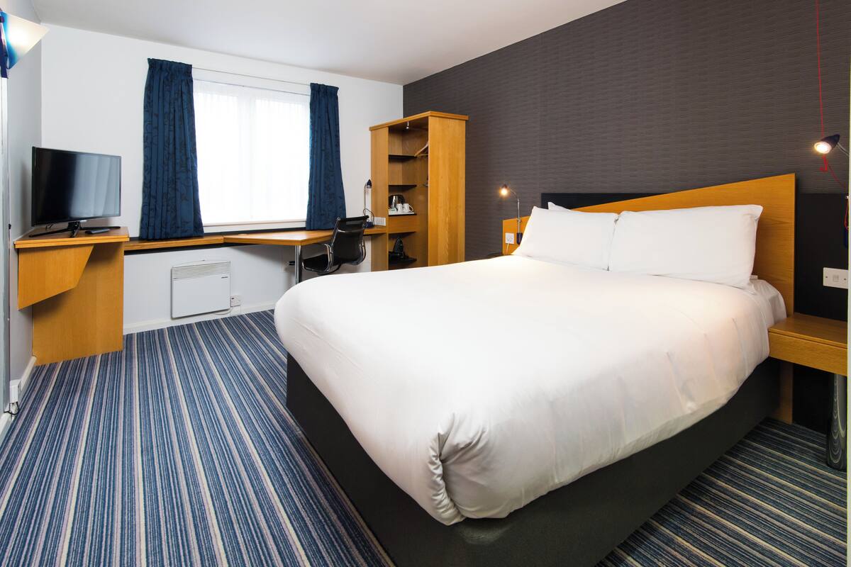 A hotel room with a large bed, a desk with a chair, a TV, a wardrobe, and a window with curtains.