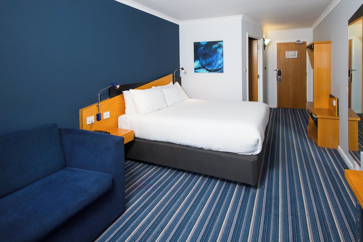 Una habitación de hotel con una cama, un sofá azul, una mesita de noche de madera, una lámpara de pared y una puerta.