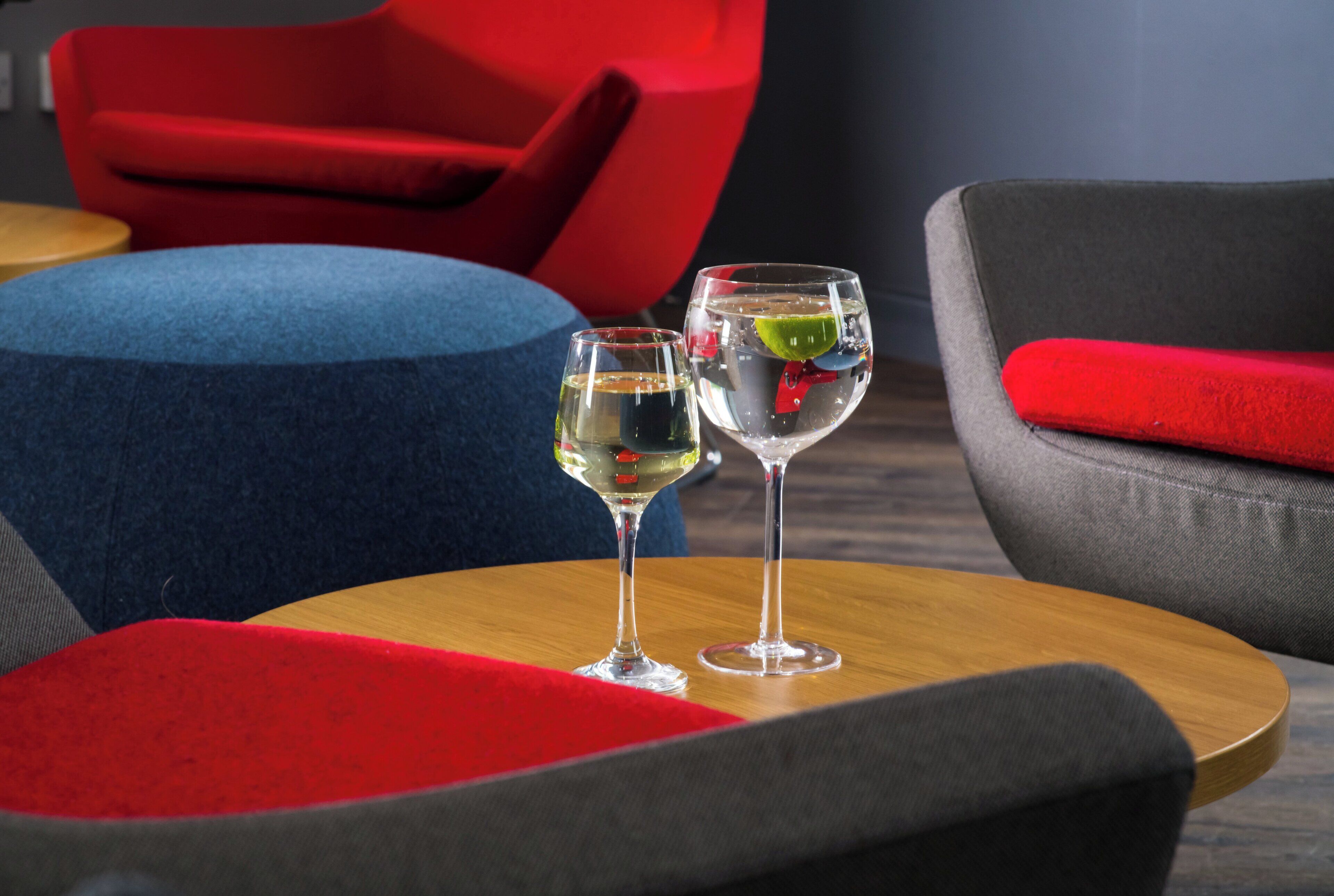 Dos vasos de un líquido transparente sobre una mesa de madera, con un cojín rojo y una silla azul al fondo.