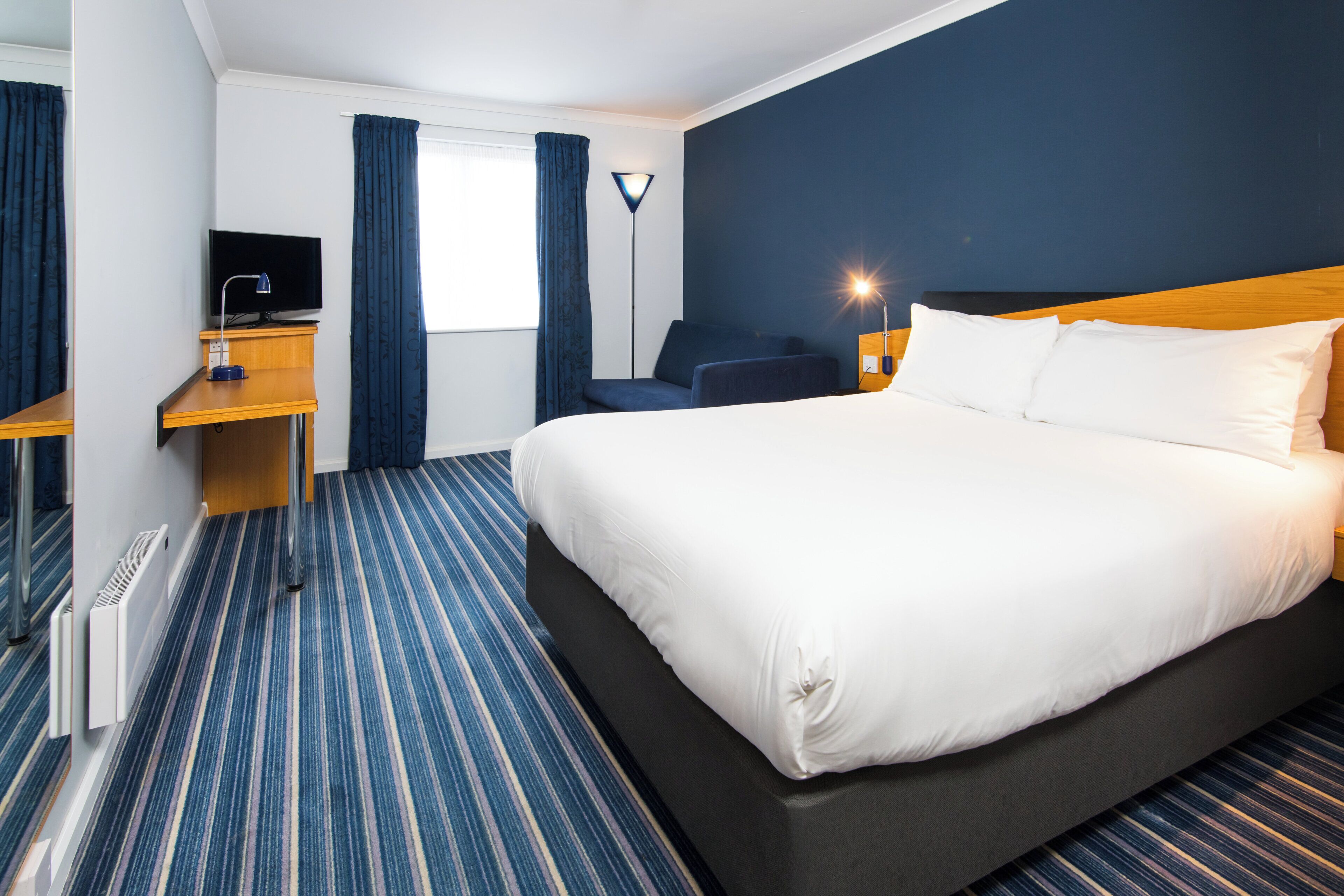 A hotel room with a large bed, a desk with a computer, a chair, and a lamp.