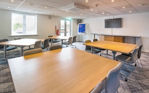 A conference room with tables and chairs, a clock, a fire extinguisher, a door, and a mounted screen. - Holiday Inn Express Manchester East by IHG (Manchester)