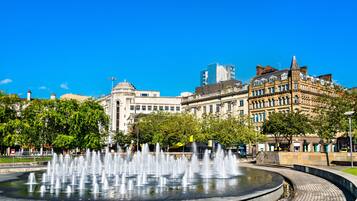 A public fountain with water jets, surrounded by historic buildings and trees.