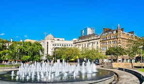 Una fuente pública con chorros de agua, rodeada de edificios históricos y árboles.