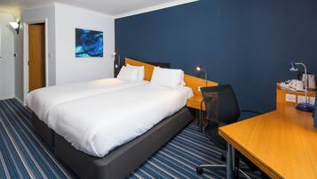 Quarto de hotel com cama, escrivaninha, cadeira, luminária de mesa, uma obra de arte na parede e porta.