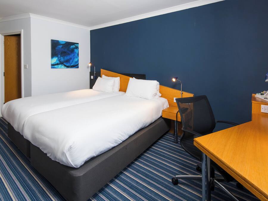 Quarto de hotel com cama, escrivaninha, cadeira, luminária de mesa, uma obra de arte na parede e porta.