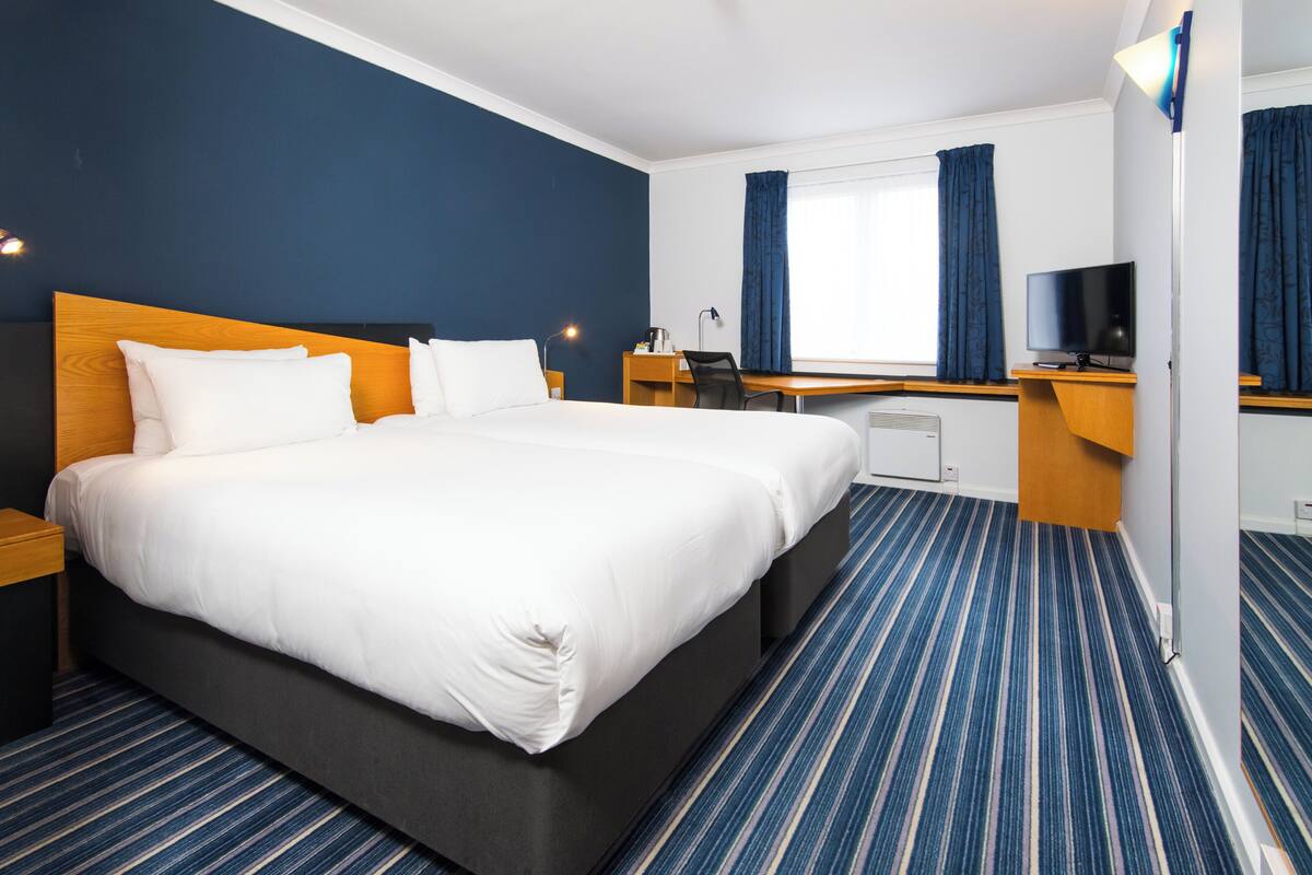 A hotel room with a large bed, a desk with a chair, a television, and a window with curtains.