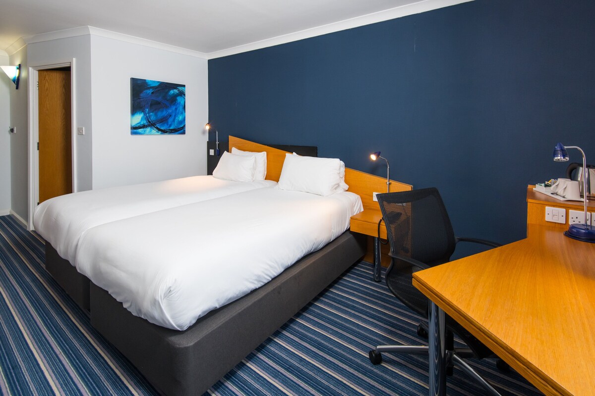 Habitación de hotel con una cama grande, un escritorio de madera, una silla, una lámpara de escritorio y un cuadro en la pared.