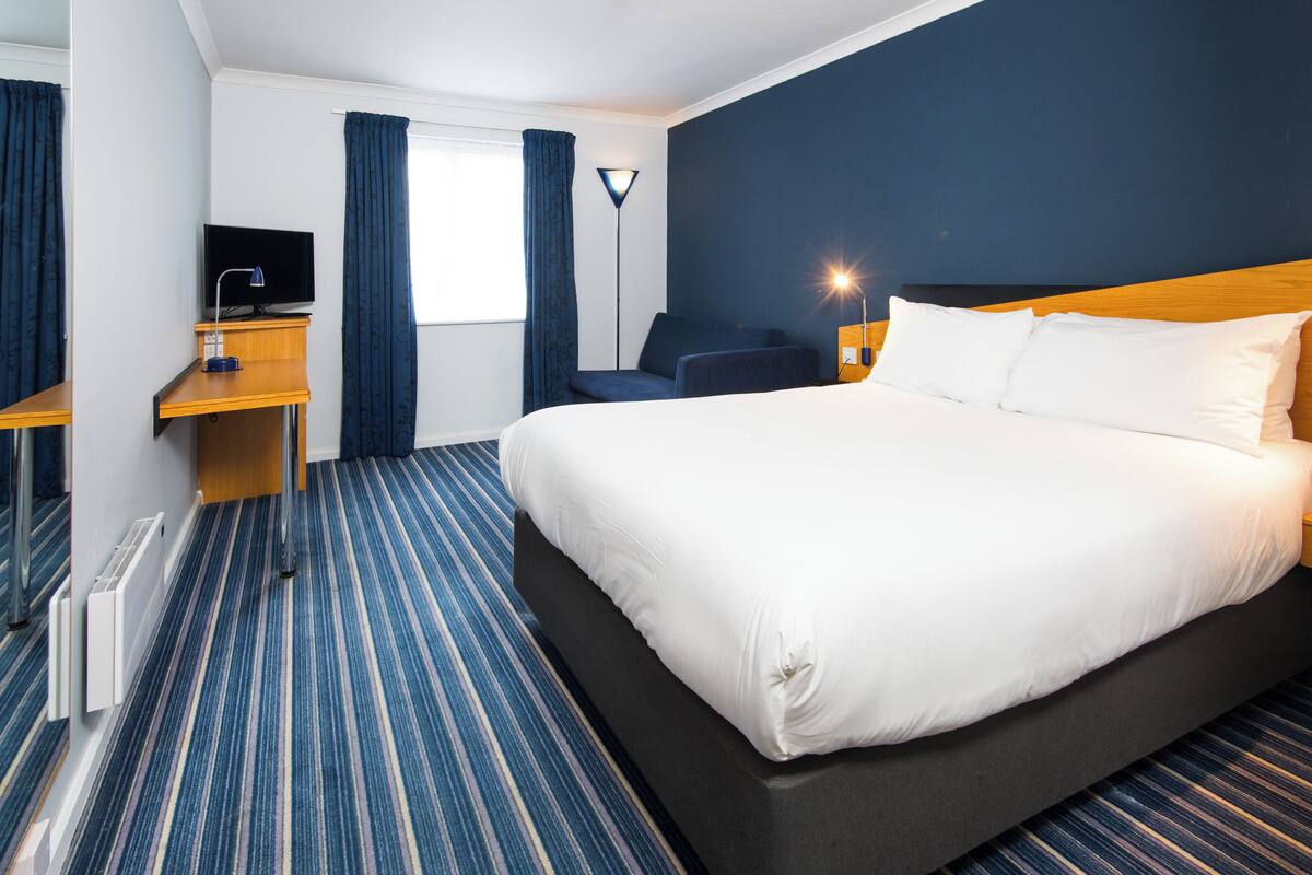 A hotel room with a large bed, a desk, a chair, a TV, and a lamp.